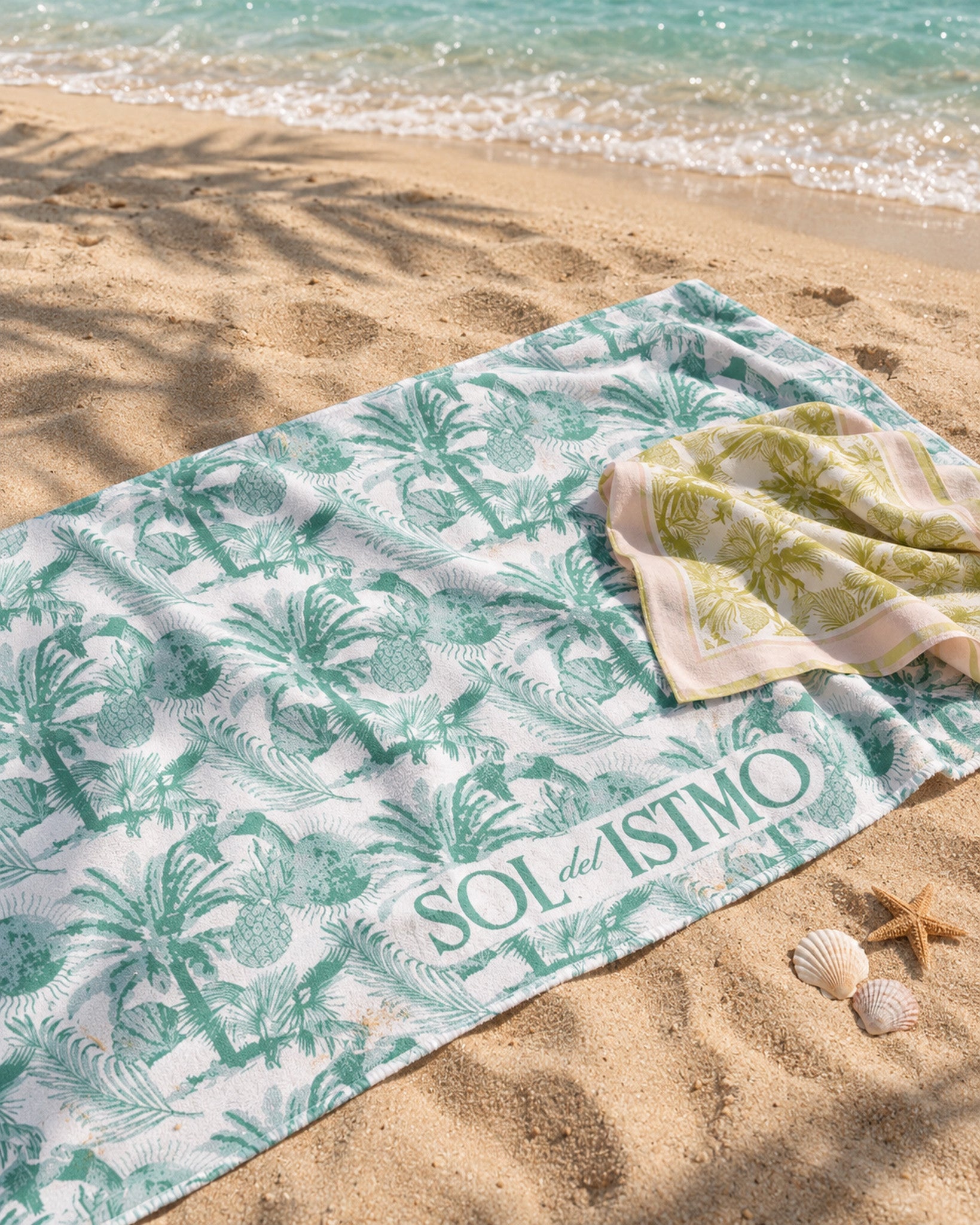 Sol del Istmo palm-print beach towel and bandana laid out on the sand at a Panamanian beach