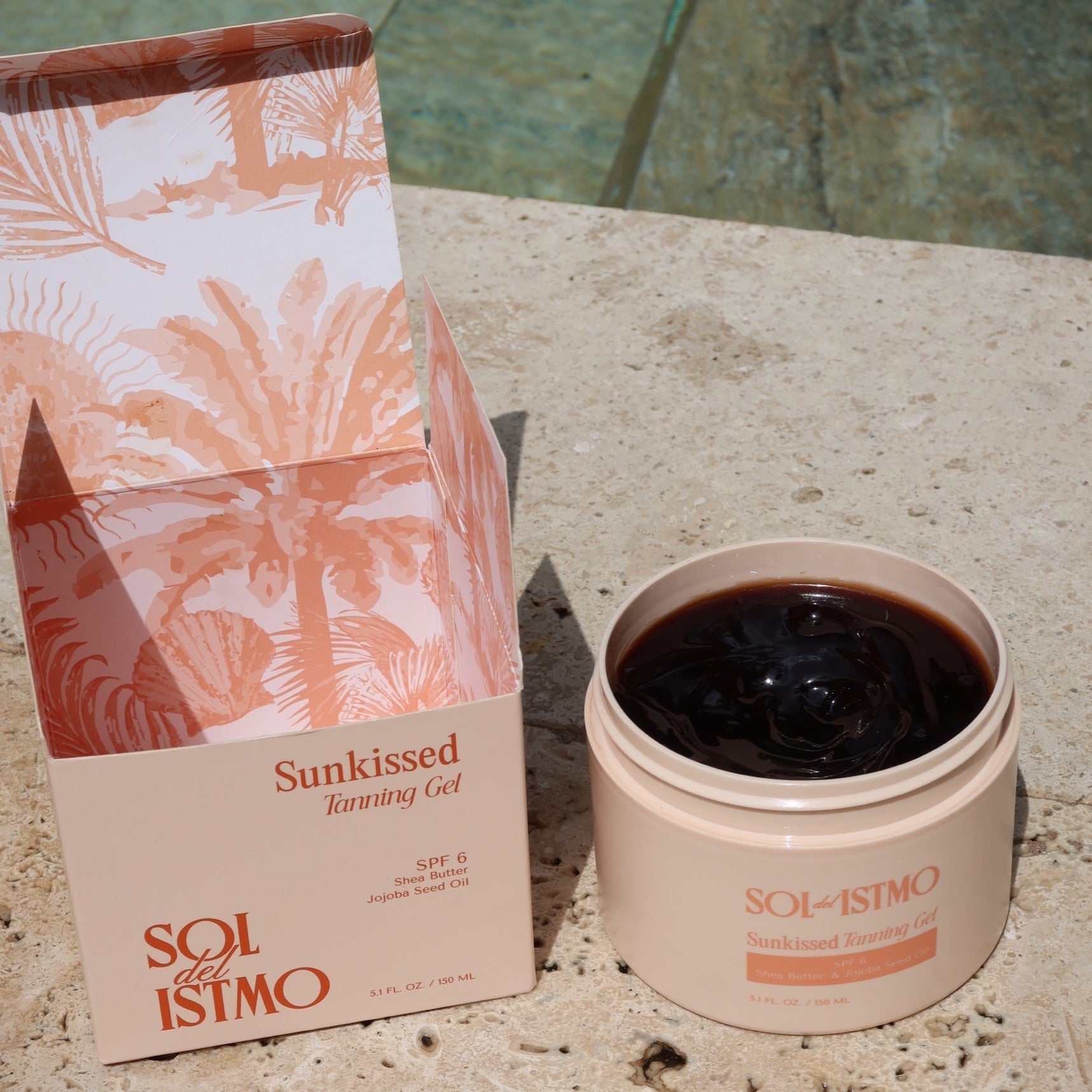 Sunkissed Tanning Gel open jar next to its palm-print box on a travertine pool edge